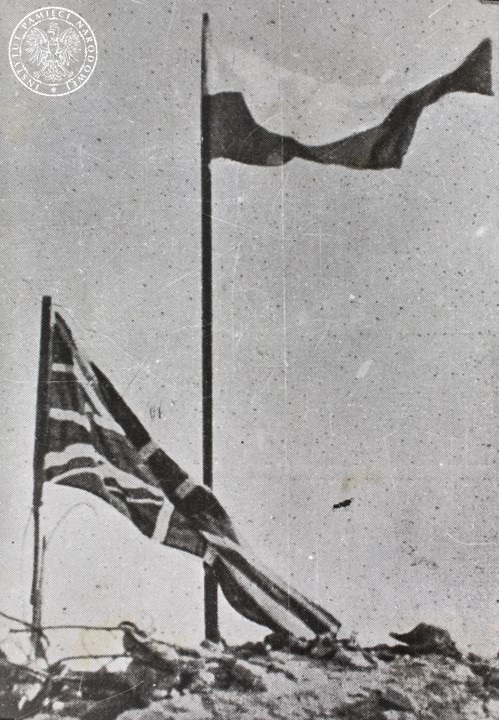 Flaga polska i brytyjska zatknięte na ruinach klasztoru Benedyktynów na Monte Cassino. Nad i pod zdjęciem napis: Polacy pierwsi w wyścigu krwi. Flaga polska zatwierdzona na szczycie „Monte Cassino” Fot. z zasobu AIPN