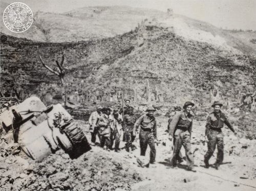 Polish soldiers carry and lead the wounded. Monte Cassino abbey is visible in the background. A damaged M4 Sherman hangs on the side of the road on the left.