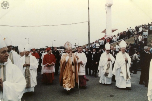 Jan Paweł II w złotej kapie i mitrze, z papieskim pastorałem w lewej ręce, idzie wzdłuż Stadionu Dziesięciolecia w Warszawie. Na lewo od niego prymas Józef Glemp w czerwonej szacie i birecie. Na prawo od papieża mistrz papieskich ceremonii liturgicznych ksiądz John Magee. Warszawa, 17 czerwca 1983 roku