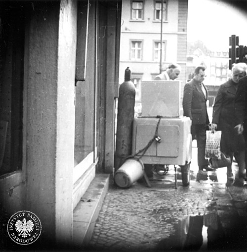 Fotografia operacyjna wykonana z ukrytego aparatu. Na pierwszym planie nieczynny, zabezpieczony przed kradzieżą, saturator. Róg ulicy Dworcowej i 15 Grudnia (obecnie Św. Jana) w Katowicach, b.d. (fot. z zasobu AIPN)