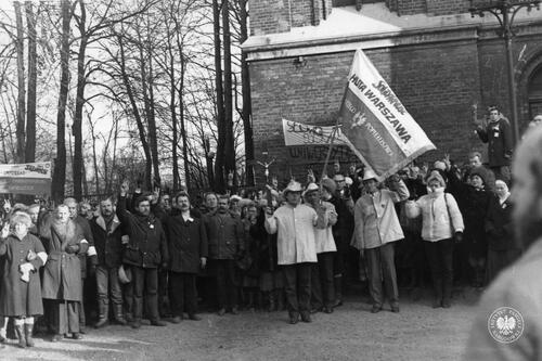 Na fotografii widać ludzi zgromadzonych pod kościołem, w tym reprezentację hutników z Huty Warszawa z zakładowym sztandarem "Solidarności". Część z uczestników uroczystości układa palce w literę "V". (fot. AIPN)