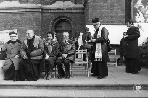 Fotografia wykonana przed kościołem, na krzesłach widać siedzącą rodzinę ks. Jerzego Popiełuszki – ojca Władysława i siostrę ks. Jerzego. Po prawej stronie, z założoną stułą, stoi o. Stefan Miecznikowski (jezuita, kapelan łódzkiej "Solidarności"). (fot. AIPN)
