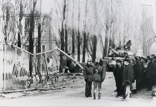 vStrajk w Stoczni Gdańskiej im. Lenina w grudniu 1981 r. Brama nr 2 Stoczni Gdańskiej im. Lenina. W centralnej części fotografii otwarta brama stoczni z widocznym zniszczonym skrzydłem. Pod budynkiem przy bramie widać szpaler ludzi. Przy nich dwaj milicjanci w hełmach, jeden z przezroczystą przyłbicą. Na budynku zerwany, zwisający fragment poszycia. Na dalszym planie widoczny czołg T55 stojący przodem w kierunku wylotu bramy