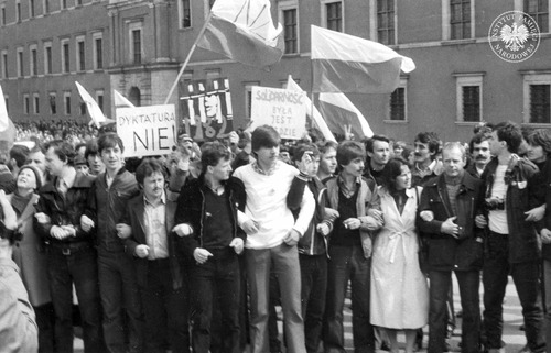 Obchody Święta Pracy zorganizowane przez podziemne struktury Niezależnego Samorządnego Związku Zawodowego „Solidarność”. Demonstranci na placu Zamkowym trzymają flagi i transparenty NSZZ „Solidarność” oraz Niezależnego Zrzeszenia Studentów (NZS). Na transparentach hasła: „SOLIDARNOŚĆ BYŁA JEST BĘDZIE”, „DYKTATURA NIE”. Warszawa, 1 maja 1982 r. (fot. z zasobu IPN)
