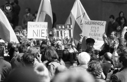 Obchody Święta Pracy zorganizowane przez podziemne struktury Niezależnego Samorządnego Związku Zawodowego „Solidarność”. Demonstranci na placu Zamkowym trzymają flagi i transparenty NZS oraz NSZZ „Solidarność” z hasłami: „SOLIDARNOŚĆ BYŁA JEST BĘDZIE” i „DYKTATURA NIE”. Warszawa, 1 maja 1982 r. (fot. z zasobu IPN)