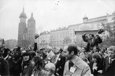 Lech Wałęsa niesiony na ramionach przez dwóch mężczyzn wśród tłumu ludzi na Rynku Głównym w Krakowie. W tłumie idą: Mieczysław Gil (po lewej z podniesiona ręką), Alina Pieńkowska (druga z prawej). W tle po prawej kościół Mariacki, Kraków 19 października 1980 roku