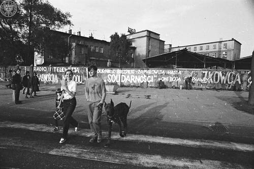 Piesi z psem na przejściu, w głębi hasła na ogrodzeniu w alei Grunwaldzkiej w Gdańsku („Radio i telewizja własnością narodu”, „Durnie z bezpieki - zamalowują hasła »Solidarności« - oni boją się prawdy i własnego cienia”). Fot. Stanisław Składanowski, 1981 r. (fot. z zasobu AIPN)