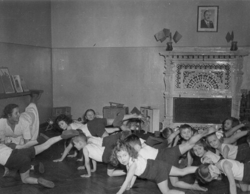 Gromada dzieci w wieku przedszkolnym wykonuje ćwiczenia gimnastyczne. W tle na ścianie wisi portret Bolesława Bieruta (fot. z zasobu IPN)
