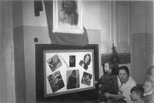 Grupa dzieci w wieku przedszkolnym słucha pogadanki swojej opiekunki. Na tablicy wisi gazetka przedstawiająca rolę kobiety w PRL-u. Nad nią portret Józefa Stalina (fot. z zasobu IPN)