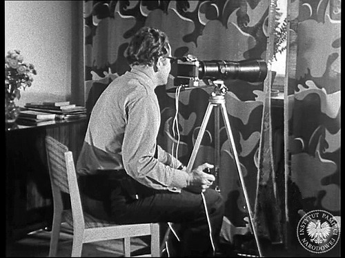 Zdjęcie funkcjonariusza w trakcie obserwacji. Kadr z filmu szkoleniowego pt. „Pentacon Super”. Film, zrealizowany na potrzeby Ośrodka Nauczania Obserwacji Ministerstwa Spraw Wewnętrznych, przedstawia opis obsługi aparatu fotograficznego Pentacon Super wraz z dodatkowym wyposażeniem oraz techniki i sposoby wykonywania przez wywiadowców zdjęć z ruchomych i stałych punktów zakrytych (z zasobu IPN)