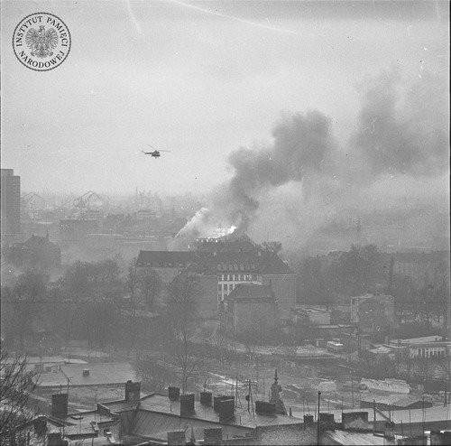 Protesty na Wybrzeżu w grudniu 1970 r. Centrum Gdańska w rejonie dworca PKP. Widoczna siedziba Dyrekcji Okręgu Poczty. Za nim płonący gmach Komitetu Wojewódzkiego PZPR. Po lewej lecący Mi-2 (fot. z zasobu AIPN)