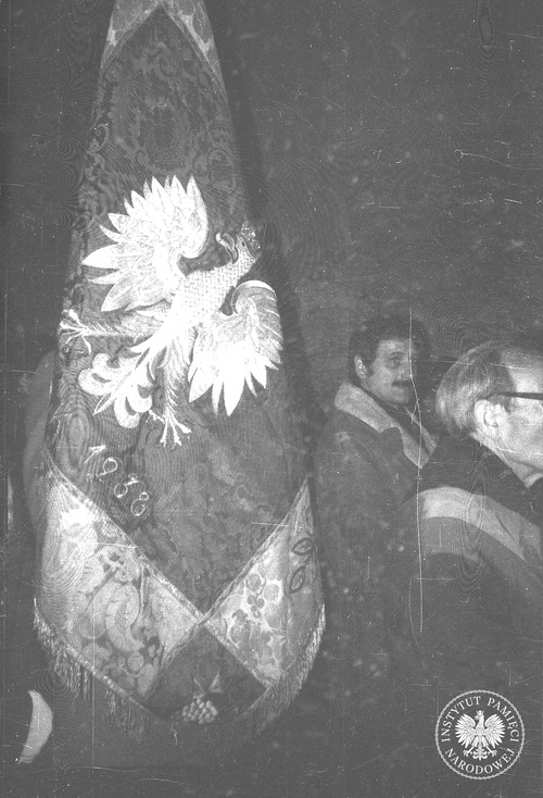 Uczestnicy nocnego zgromadzenia przed Grobem Nieznanego Żołnierza na pl. Zwycięstwa (obecnie pl. Piłsudskiego) w Warszawie z okazji nieakceptowanego przez władze PRL Święta Niepodległości. Widoczni mężczyźni z sztandarem, na którym wyhaftowany jest Orzeł Biały w koronie. 1979–1983. Fot. Jerzy Lech Szóstko, Edward Kucharski, z zasobu AIPN