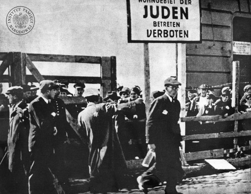 Żydzi oczekujący na przejście przez bramę łódzkiego getta przy ul. Zgierskiej, należącej do części tzw. aryjskiej, u wylotu ul. Lutomierskiej. U góry tablica z niemieckim napisem: „[Wohngebiet] der Juden. Betreten verboten” (Żydowska dzielnica mieszkaniowa. Wstęp wzbroniony). Pośrodku funkcjonariusz żydowskiej Służby Porządkowej. Łódź, 1940 r. (fot. z zasobu AIPN)
