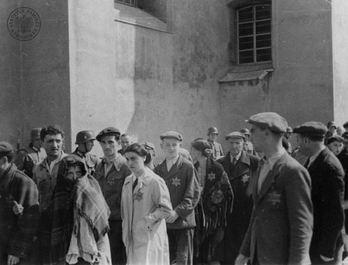Grupa Żydów prowadzona ulicą łódzkiego getta. Widoczne gwiazdy naszyte na ubraniach. Z tyłu stoją niemieccy policjanci z karabinami. Łódź, 1940–1944 r. (fot. z zasobu AIPN)