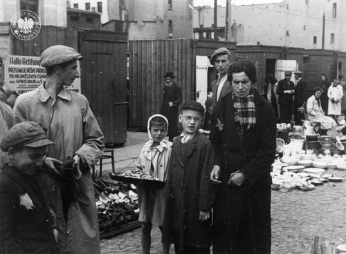 Mieszkańcy łódzkiego getta na targowisku Jojny Pilcera przy ul. Łagiewnickiej. Na pierwszym planie widoczne m.in. handlujące dziecko. Z tyłu stoiska z rozłożonym towarem – butami i naczyniami. Łódź, 1940–1941 r. (fot. z zasobu AIPN)