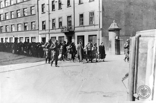 Wysiedlenie Żydów z getta krakowskiego do obozu płaszowskiego. Żydowska ludność Krakowa, niosąc swój dobytek, pod eskortą SS-manów opuszcza teren getta. Na ostatnim planie kamienice stojące przy ulicy Lwowskiej. Marzec 1943 r. (fot. z zasobu AIPN)