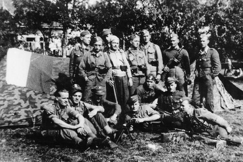 Partyzanci OP „Żelbet I” (20. pp AK) w Komornikach koło Raciechowic, przełom września i października 1944 r. Nad namiotem polska flaga, nieformalny sztandar oddziału. Widoczny liczny niemiecki ekwipunek, m.in. peleryny maskujące, zdobyte podczas akcji w Tokarni. Fot. ze zbiorów P. Sadowskiego