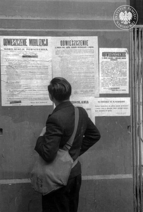Młody mężczyzna z amerykańską torbą wojskową na ramieniu czyta obwieszczenie dotyczące mobilizacji powszechnej. Charakterystyczne na tym afiszu są dwie ciemne pieczęcie zamazujące poprzednią datę rozpoczęcia mobilizacji. Dodatkowo po lewej m.in. plakat: „Obwieszczenie o poborze koni, wozów, pojazdów mechanicznych i rowerów”. Warszawa, wrzesień 1939 r. Skan fotografii z Kolekcji Juliena Bryana przechowywanej w USHMM w Waszyngtonie (fot. z zasobu AIPN)